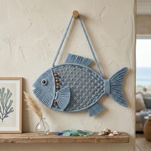 May include: A blue macrame fish wall hanging featuring a knotted cord body, a single dark eye, and wooden bead accents. The fish has a braided cord loop for hanging and is constructed with fringe details on the dorsal, pectoral, and tail fins.
