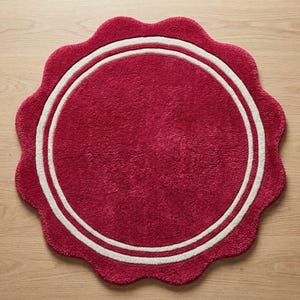May include: A circular rug with a deep red, textured pile featuring a scalloped edge and two concentric white rings near the perimeter. The rug has a soft, tufted appearance and maintains a consistent color throughout the central circular area.