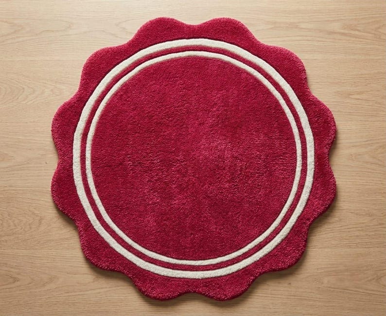 May include: A circular rug with a deep red, textured pile featuring a scalloped edge and two concentric white rings near the perimeter. The rug has a soft, tufted appearance and maintains a consistent color throughout the central circular area.