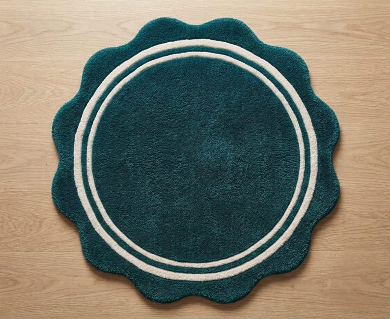 May include: A round, teal-colored rug with a scalloped edge and a double white circular border. The rug features a plush, textured surface and a consistent deep teal hue throughout the center, framed by two concentric white rings near the perimeter.