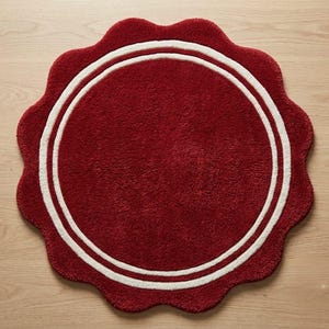 May include: A circular rug with a deep red, textured surface and a scalloped edge. The rug features two concentric white circular lines that frame the center, creating a distinct border pattern against the solid red background of the textile.