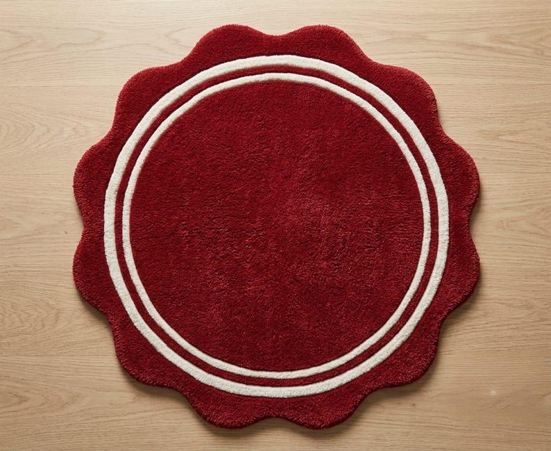May include: A circular rug with a deep red, textured surface and a scalloped edge. The rug features two concentric white circular lines that frame the center, creating a distinct border pattern against the solid red background of the textile.