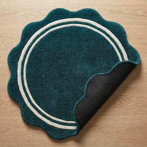 May include: A circular teal rug with a scalloped edge and two concentric white rings. The rug features a plush, tufted texture on the top surface, while one corner is folded back to reveal a solid black, non-slip backing material underneath.
