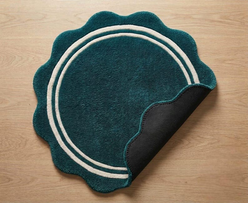 May include: A circular teal rug with a scalloped edge and two concentric white rings. The rug features a plush, tufted texture on the top surface, while one corner is folded back to reveal a solid black, non-slip backing material underneath.