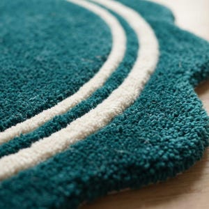 May include: A teal rug with a scalloped edge and two concentric white circular lines. The rug features a thick, tufted texture and a rounded, decorative border design that creates a distinct shape against the flat surface underneath it.