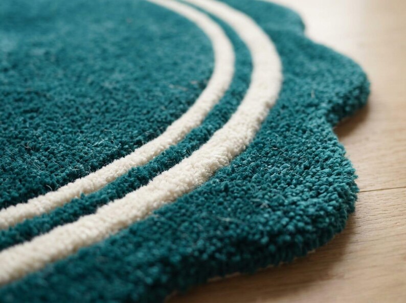 May include: A teal rug with a scalloped edge and two concentric white circular lines. The rug features a thick, tufted texture and a rounded, decorative border design that creates a distinct shape against the flat surface underneath it.