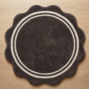 May include: A circular rug with a scalloped edge, featuring a dark charcoal gray center and a thick, cream-colored circular border. The rug has a textured, plush pile surface and is presented in a flat, top-down view on a light wood surface.