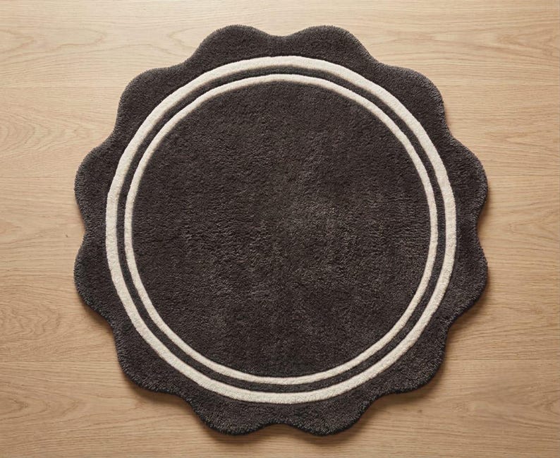 May include: A circular rug with a scalloped edge, featuring a dark charcoal gray center and a thick, cream-colored circular border. The rug has a textured, plush pile surface and is presented in a flat, top-down view on a light wood surface.