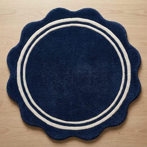 May include: A circular rug with a scalloped edge, featuring a deep navy blue color and two concentric white rings near the perimeter. The rug has a thick, tufted texture throughout its surface, creating a soft and plush appearance for floor decor.