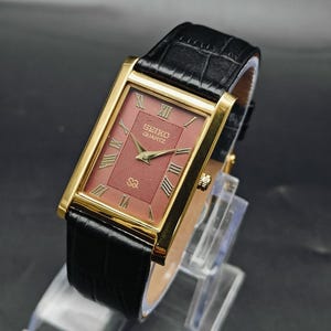 May include: A rectangular Seiko Quartz wristwatch featuring a gold-tone case and a textured maroon dial with gold Roman numeral hour markers. The watch includes gold-tone hands and a black leather strap with a crocodile-embossed pattern.