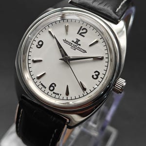 Op de afbeelding: Een roestvrijstalen polshorloge met een witte wijzerplaat, zwarte Arabische cijfers op 3, 6, 9 en 12, en zilverkleurige wijzers. De wijzerplaat bevat de tekst Jaeger-LeCoultre Club en T Swiss Made T. Zwarte lederen band met witte stiksels.