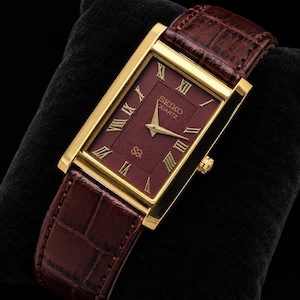May include: A rectangular Seiko Quartz watch with a gold-tone case and a deep burgundy dial featuring a textured wave pattern. The watch has gold Roman numeral hour markers, gold hands, and a brown leather strap with a crocodile-embossed texture.