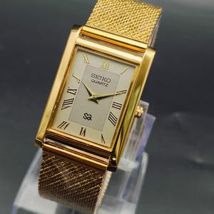 Op de afbeelding: Een rechthoekig Seiko Quartz polshorloge in goudkleur met een crèmekleurige wijzerplaat, Romeinse cijfers, een gestructureerd middenpatroon en een goudkleurige metalen mesh-band. De wijzerplaat toont het Seiko Quartz logo en een klein embleem.