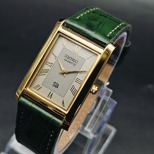 Op de afbeelding: Een rechthoekig Seiko Quartz polshorloge met een goudkleurige kast en een donkergroene leren band. De wijzerplaat heeft een zilverkleurig midden met een golvend lijnpatroon, gouden Romeinse cijfers, gouden wijzers en het Seiko-logo.