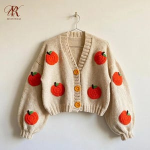 Könnte beinhalten: Eine beigefarbene Strickjacke mit V-Ausschnitt, langen Ärmeln mit gerippten Bündchen und drei orangefarbenen Knöpfen. Das Kleidungsstück ist mit mehreren gehäkelten orangefarbenen Kürbismotiven verziert, die jeweils einen kleinen grünen Stiel haben und über die Vorderseite und die Ärmel verteilt sind.