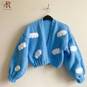 Könnte beinhalten: Eine hellblaue Strickjacke mit V-Ausschnitt, langen Ärmeln mit gerippten Bündchen und einem gerippten Saum. Das Kleidungsstück ist mit mehreren erhabenen, weißen, strukturierten, wolkenförmigen Applikationen auf der Vorderseite und den Ärmeln verziert.