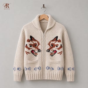 Könnte beinhalten: Eine cremefarbene Strickjacke mit Reißverschluss, Schalkragen und zwei Vordertaschen. Das Kleidungsstück zeigt zwei symmetrische Fuchskopfmotive in Braun, Schwarz und Rot sowie eine Reihe kleiner blauer Pfotenabdrücke am unteren Saum.
