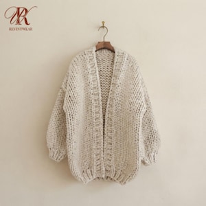 May include: An open-front cardigan sweater in a chunky, cream-colored knit with subtle brown flecks. The garment features long sleeves, a ribbed hem, and a vertical ribbed placket along the front opening. The logo REVINTWEAR is visible in the corner.
