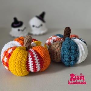 May include: Two crocheted pumpkins with brown stems sit in the foreground. The left pumpkin features orange, yellow, red, and white segments. The right pumpkin has blue, brown, and white sections. Two small white crocheted ghosts are in the background.