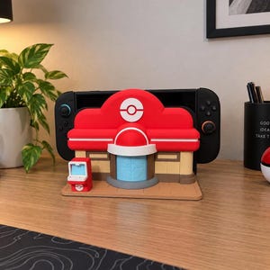 May include: A red, white and tan plastic gaming console dock shaped like a building with a circular emblem on top. The structure features a light blue door, a small red side console and a flat base, designed to hold a black handheld gaming device.
