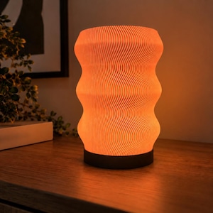 May include: A cylindrical table lamp with a textured, wavy exterior surface that emits a warm, orange-toned glow. The lamp features a repeating vertical pattern of fine, undulating lines and is supported by a solid, dark-coloured circular base.