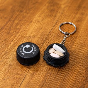 May include: A black circular pill container keychain with a screw-on lid featuring a white recycling symbol and the text RECYCLE ME. The open container holds several oval-shaped pills in shades of white and beige, attached to a metal ring.