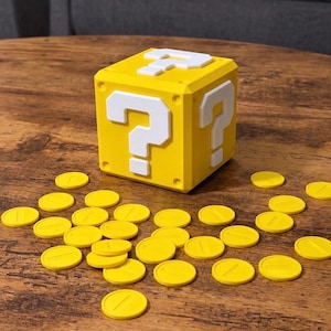 Op de afbeelding: Een gele kubus met een verhoogd wit vraagteken op elke zichtbare zijde, vergezeld door een verzameling kleine, platte, ronde gele penningen die rond de basis van de kubus op een houten oppervlak verspreid liggen.