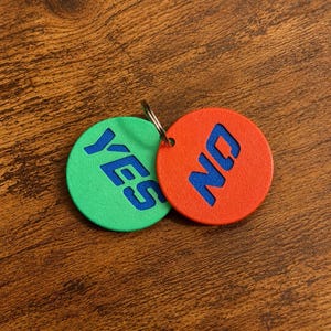 May include: Two circular tokens attached to a metal ring. One token is green with the word YES embossed in blue, and the other is orange with the word NO embossed in blue. The tokens have a textured surface and are positioned side by side.