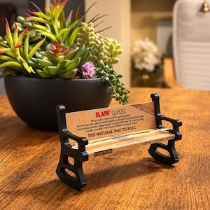 May include: A miniature black plastic bench designed to hold rolling papers, featuring the text RAW CLASSIC, We proudly celebrate 20 years of RAW!, THE NATURAL WAY TO ROLL, 32 LEAVES PER PACK, 32 CARTINE, and 32 FOLMS printed on the backrest and seat.