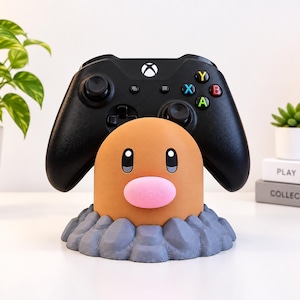 May include: A black Xbox game controller resting on a tan, mole-like character stand with a pink nose and dark eyes, set upon a textured grey base that resembles rocky ground. The character design features a smooth, rounded top surface.