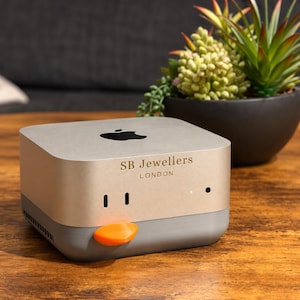 May include: A square, metallic gold and gray electronic device featuring a black Apple logo on the top surface. The front panel displays the text SB Jewellers LONDON in black lettering, with two vertical slots, a small light, and an orange tab.
