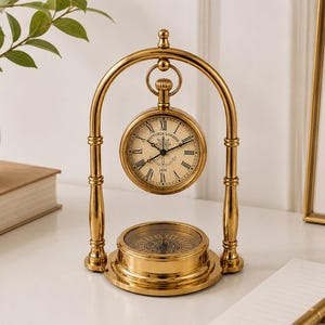 May include: A gold-toned metal desk clock and compass set featuring an arched stand. The clock face displays Roman numerals, the text Victoria London, and a central anchor graphic. A circular compass is integrated into the base of the stand.