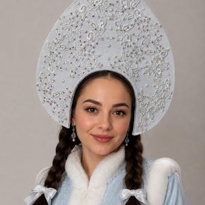 Könnte beinhalten: Ein hellblauer Kokoshnik-Kopfschmuck mit aufwendiger weißer Spitzenstickerei, verstreuten irisierenden Kristallen und kleinen weißen Perlen. Das hohe, bogenförmige Kopfstück ist mit einer spitzen Oberseite und geschwungenen Seiten gestaltet und dient als detailliertes Accessoire.