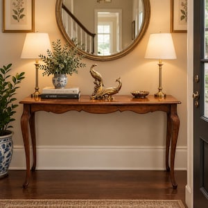 May include: A wooden console table with curved legs, topped with two brass lamps with white shades, a blue and white patterned vase with greenery, two stacked books, two gold-toned peacock figurines, and a small bowl, placed below a round mirror.