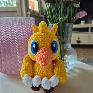Könnte beinhalten: Eine gelbe, gehäkelte Plüschvogel-Figur mit strukturierter Garnoberfläche. Die Figur hat einen markanten orangefarbenen Schnabel, große blaue und schwarze Augen, einen kleinen Kamm auf dem Kopf sowie weiße Füße mit dunkelbraunen Zehen an der Basis.