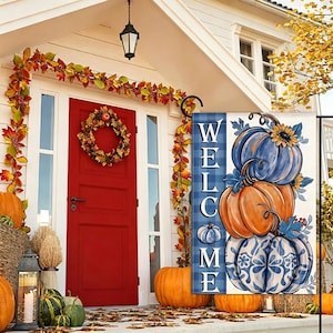 May include: A rectangular garden flag featuring a blue plaid side panel with the vertical text WELCOME in white letters. The main graphic displays a stack of three pumpkins in blue, orange, and blue-and-white floral patterns with sunflowers.