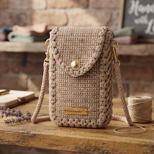 May include: A beige crochet crossbody bag featuring a textured stitch pattern, a fold-over flap secured with a gold-tone metal button, and a matching crochet strap. A rectangular gold-tone metal tag labelled "Handmade" is attached to the front.
