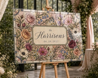 May include: A rectangular wedding sign on a wooden easel featuring a stained glass style floral border with pink, orange, and purple flowers. The center panel is cream-colored with the text "the Harrisons" and the date "06.20.2027" in black.