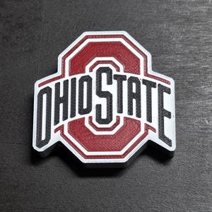 May include: A three-dimensional Ohio State University logo emblem featuring a central red block O with a white border and the word OHIO STATE in black lettering. The design has a layered, textured appearance with distinct white, red, and black sections.