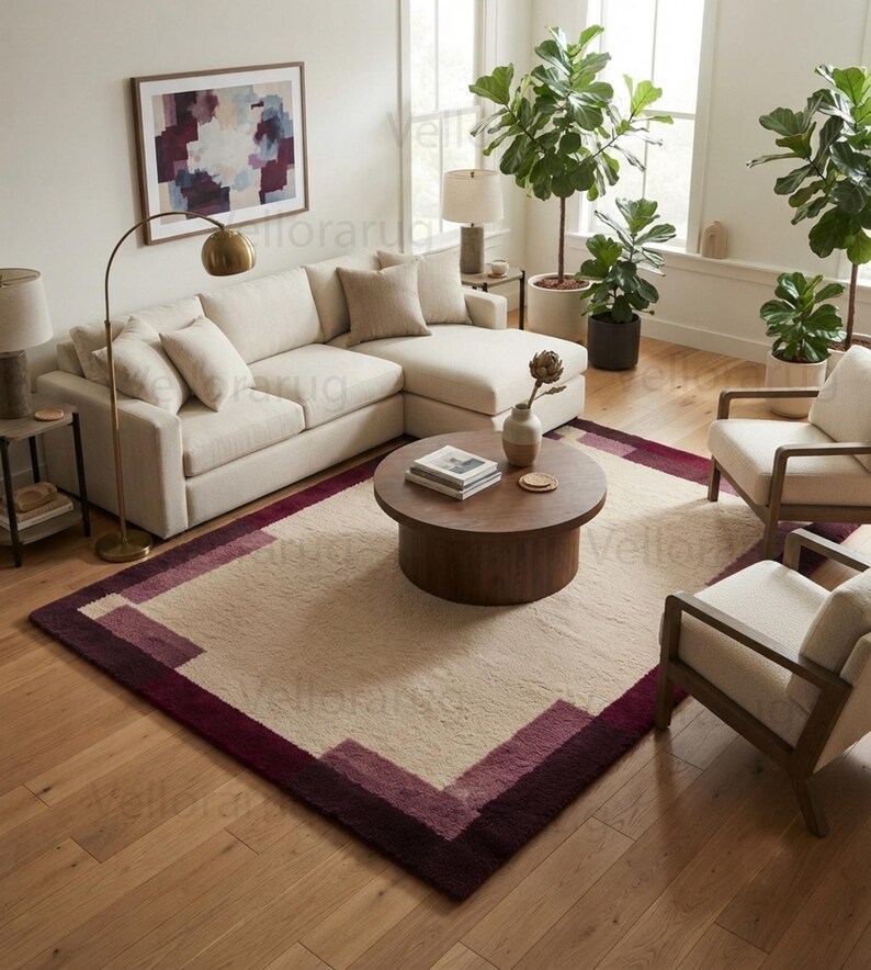 May include: A rectangular area rug featuring a large cream-colored center bordered by thick, geometric bands in shades of deep burgundy and muted plum. The rug has a textured, plush pile and is positioned on a light-toned wooden floor in a living room.