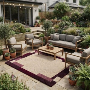 May include: A rectangular area rug featuring a central beige field framed by a deep burgundy and plum border with geometric block patterns. The rug is placed on a stone patio beneath a wooden outdoor furniture set including a sofa and chairs.