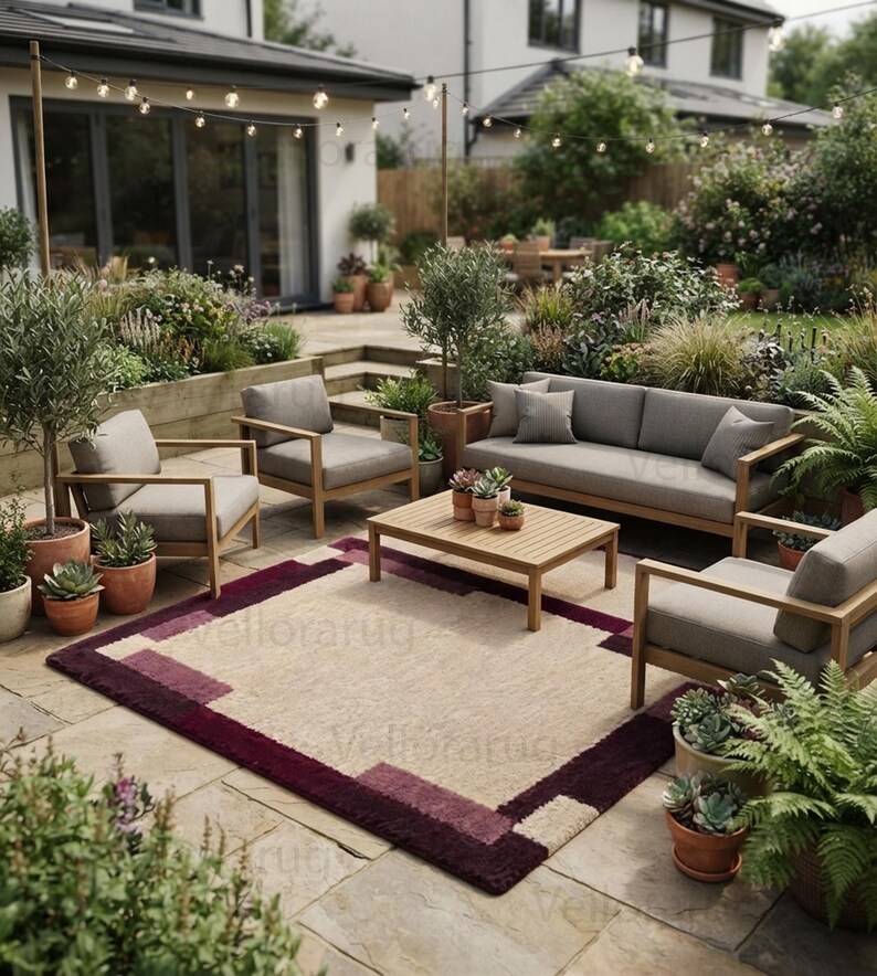May include: A rectangular area rug featuring a central beige field framed by a deep burgundy and plum border with geometric block patterns. The rug is placed on a stone patio beneath a wooden outdoor furniture set including a sofa and chairs.