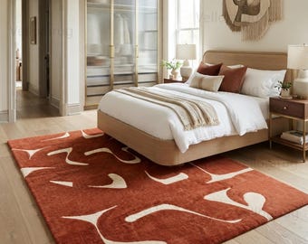 May include: A rectangular area rug featuring a deep terracotta orange base color with an abstract pattern of cream-colored, curved, organic shapes scattered across the surface. The rug is positioned on a light-toned wooden floor in a bedroom.