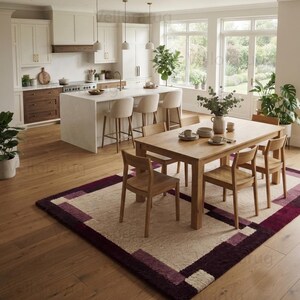 May include: A rectangular area rug featuring a geometric pattern with a cream-colored center and deep purple and plum borders. The rug is placed on a light brown wooden floor in a kitchen and dining area, positioned beneath a wooden dining table.