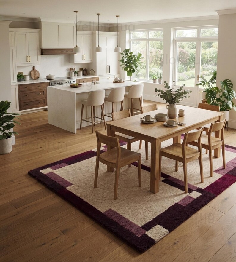 May include: A rectangular area rug featuring a geometric pattern with a cream-colored center and deep purple and plum borders. The rug is placed on a light brown wooden floor in a kitchen and dining area, positioned beneath a wooden dining table.