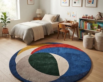 May include: A large circular area rug featuring a bold abstract design with overlapping geometric shapes in deep blue, forest green, mustard yellow, cream, and light gray. The rug has a soft, textured pile and sits on a light wood floor.