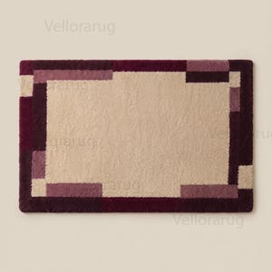 May include: A rectangular area rug featuring a large, solid beige center panel surrounded by a thick, geometric border composed of alternating blocks in deep burgundy, dark plum, and lighter mauve tones, creating a structured, layered frame effect.