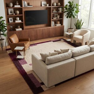 May include: A large rectangular area rug featuring a cream-colored center with a geometric border pattern in shades of deep purple and burgundy. The rug is positioned in a living room setting with a beige sofa, a brown leather chair, and a cream chair.