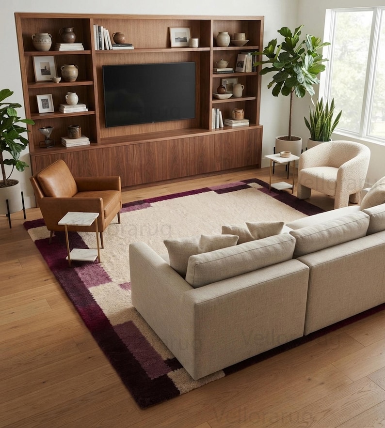 May include: A large rectangular area rug featuring a cream-colored center with a geometric border pattern in shades of deep purple and burgundy. The rug is positioned in a living room setting with a beige sofa, a brown leather chair, and a cream chair.