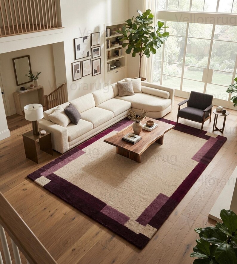 May include: A rectangular area rug featuring a large cream-colored center panel framed by a wide border in shades of deep burgundy and plum. The rug has a textured pile and is positioned on a light-toned wooden floor in a living room setting.